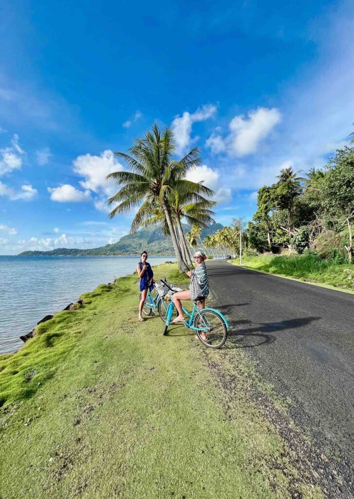 Eva et Manon à vélo au bord de l’eau à Bora Bora, avec cocotiers et montagnes en arrière-plan.