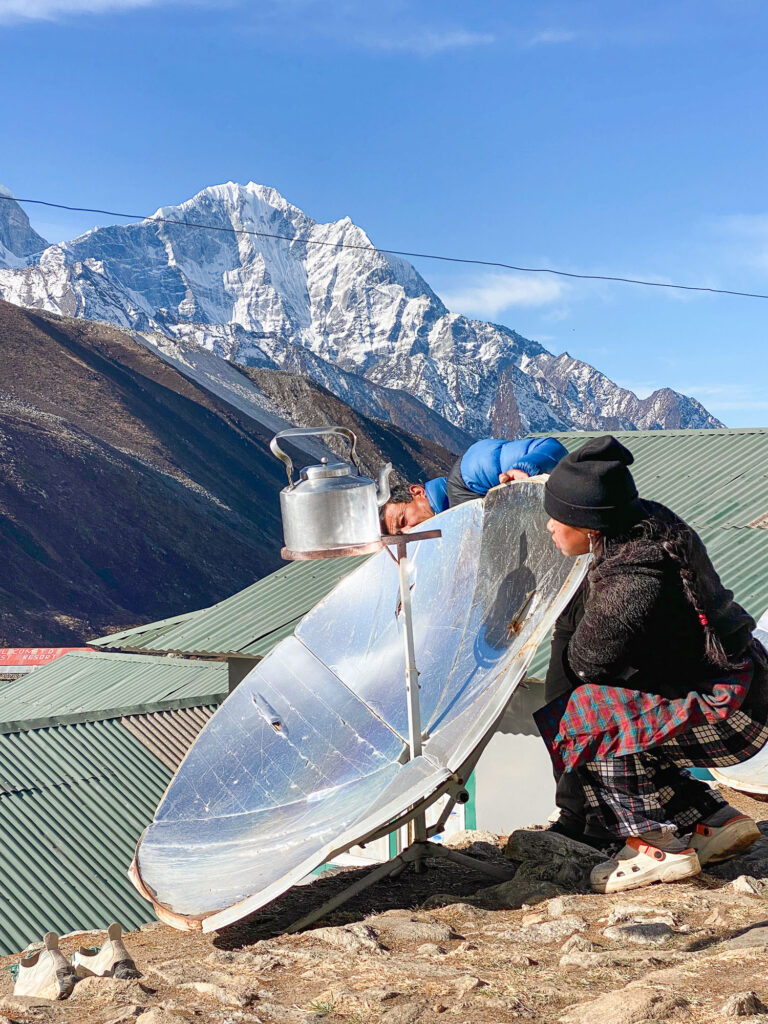 Local using a solar panel to heat water in a Nepalese village, with snow-capped Himalayan mountains in the background.