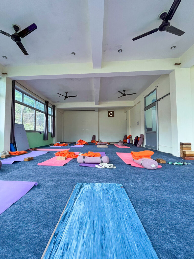 Eva sitting on her yoga mat with a view of the training hall in Rishikesh, India