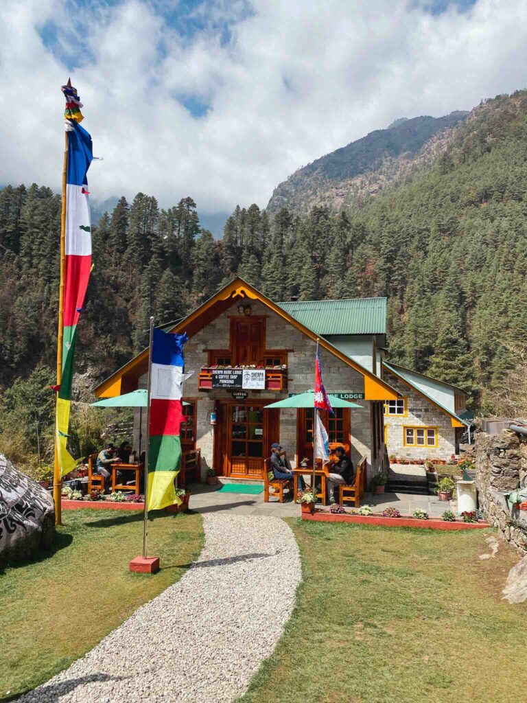 Exterior view of a traditional tea house in a Himalayan village, surrounded by green landscapes and colorful prayer flags, Three Passes Trek, Nepal.